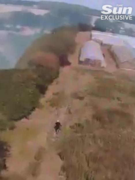 Aerial view of a person running across a rural landscape with agricultural buildings in the background.