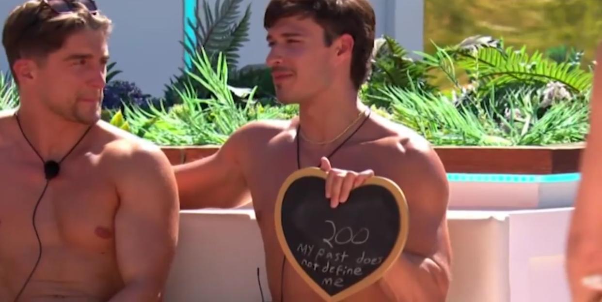 Two men are shirtless, and one holds a heart-shaped chalkboard that says "200 my past does not define me."