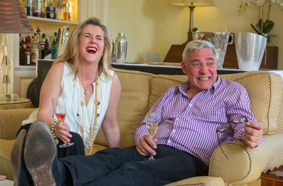 Two Gogglebox stars, a woman and a man, laugh as they sit on a couch with drinks.