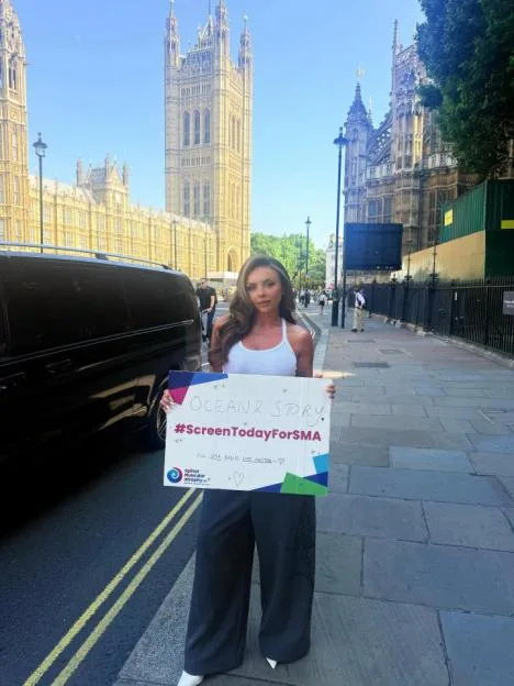 Jesy Nelson holds a sign that reads "#ScreenTodayForSMA" in front of the Palace of Westminster in London.