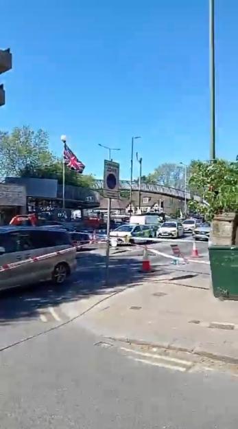 Police cars and tape blocking a street under a bright blue sky, with a Union Jack flag visible in the background.