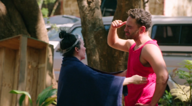 A woman wearing swimming goggles and a towel embracing a man in a red tank top.