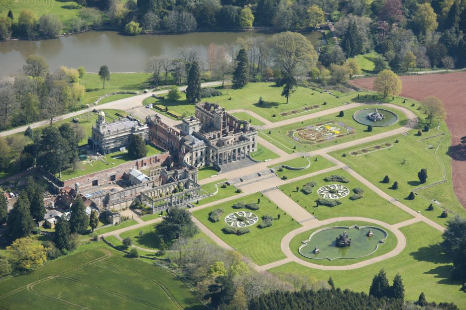 Aerial view of the ruined Witley Court mansion and surrounding landscaped gardens with a lake in the background.