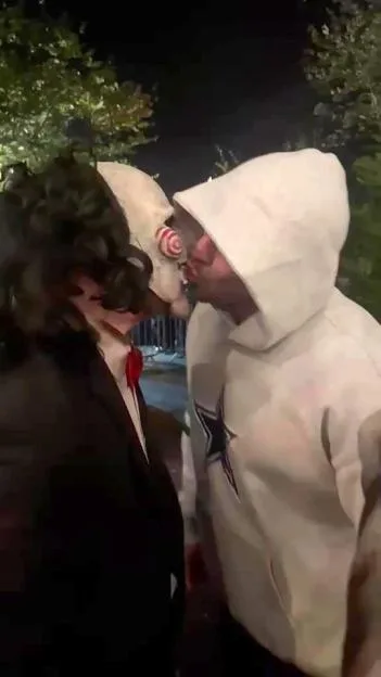 Tommy Fury in a white hoodie kissing someone wearing a Billy the Puppet mask.