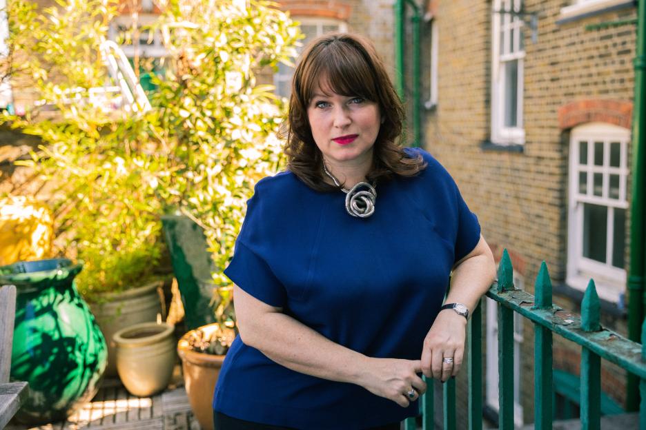Esther Stanhope, a woman with long brown hair, wearing a blue shirt and a silver statement necklace, leans on a green railing outside.