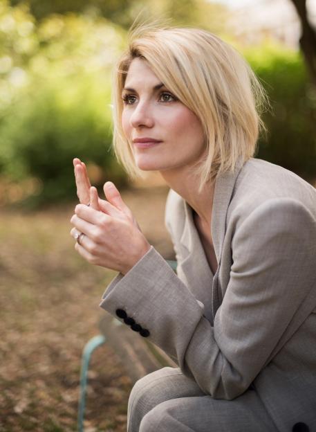 Jodie Whittaker, with short blonde hair, wearing a light gray blazer, looking into the distance with her hands near her face.