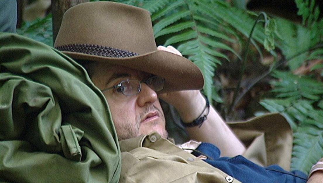 Joe Pasquale, wearing glasses and a hat, lying down in a jungle setting from "I'm a Celebrity, Get Me Out of Here!"