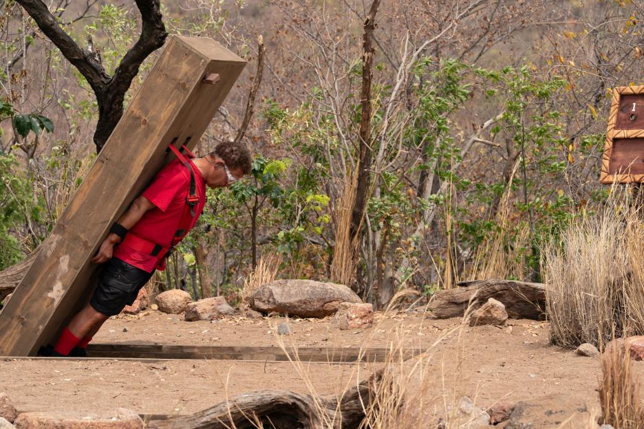 Craig Charles from "I'm A Celebrity... Get Me Out Of Here! South Africa" participating in a "Tomb Raider" challenge, suspended by a wooden contraption in a jungle setting.