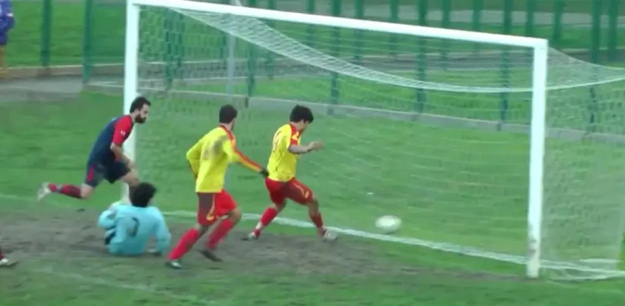A soccer player in a red and yellow jersey kicks the ball past a blue-shirted goalie, into the goal.