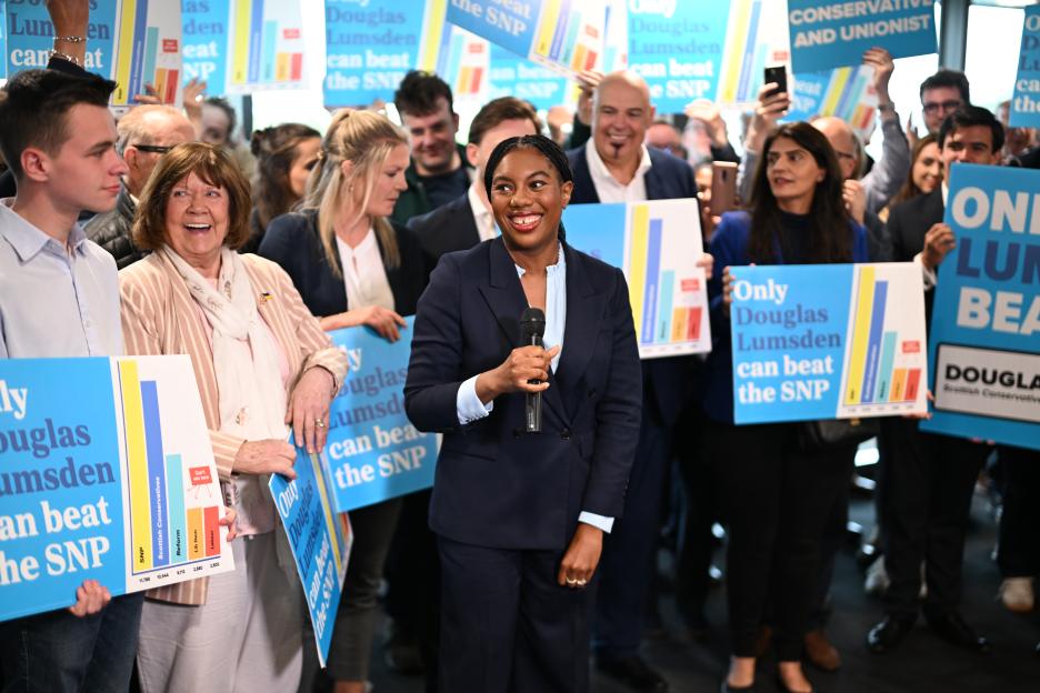 Conservative Party leader Kemi Badenoch speaking at a campaign event, surrounded by supporters holding signs for Douglas Lumsden.