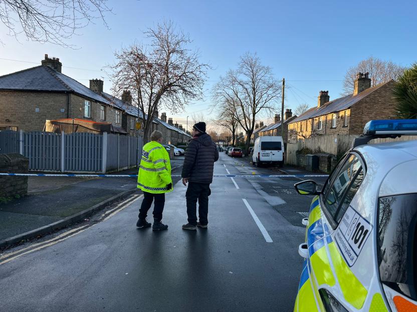 Police officer and civilian standing in a cordoned-off street with a police vehicle nearby.