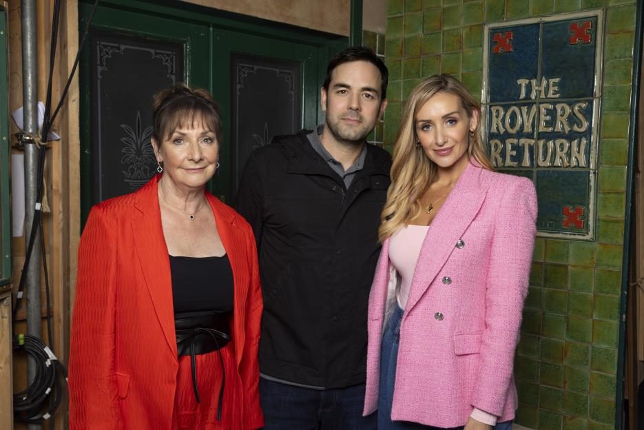 Actors Pauline McLynn, Aaron McCusker, and Catherine Tyldesley posing in front of "The Rovers Return" sign.