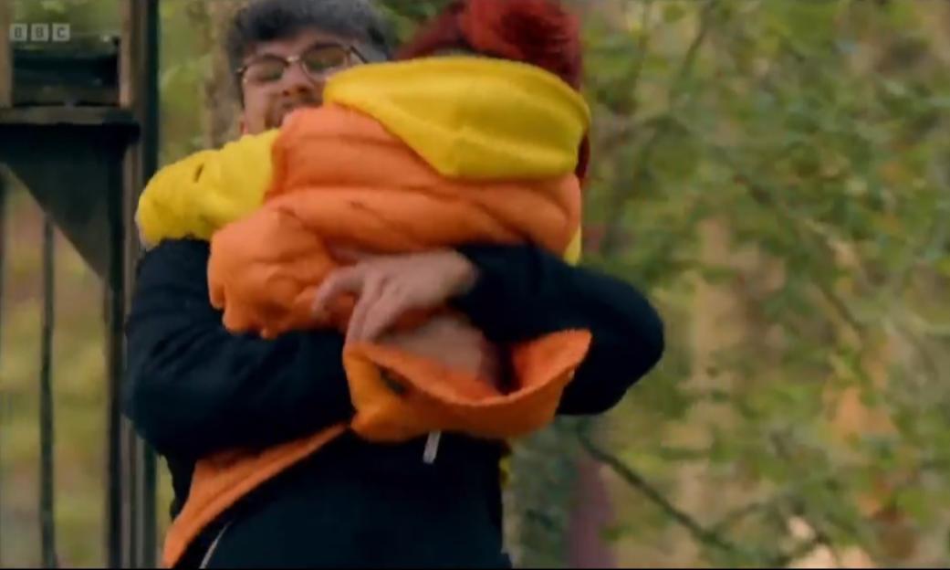 Man carrying a person wrapped in a bright orange and yellow jacket.