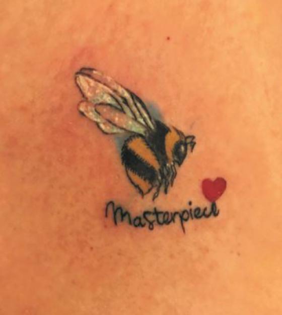 Tattoo of a bee with the word "Masterpiece" and a red heart.