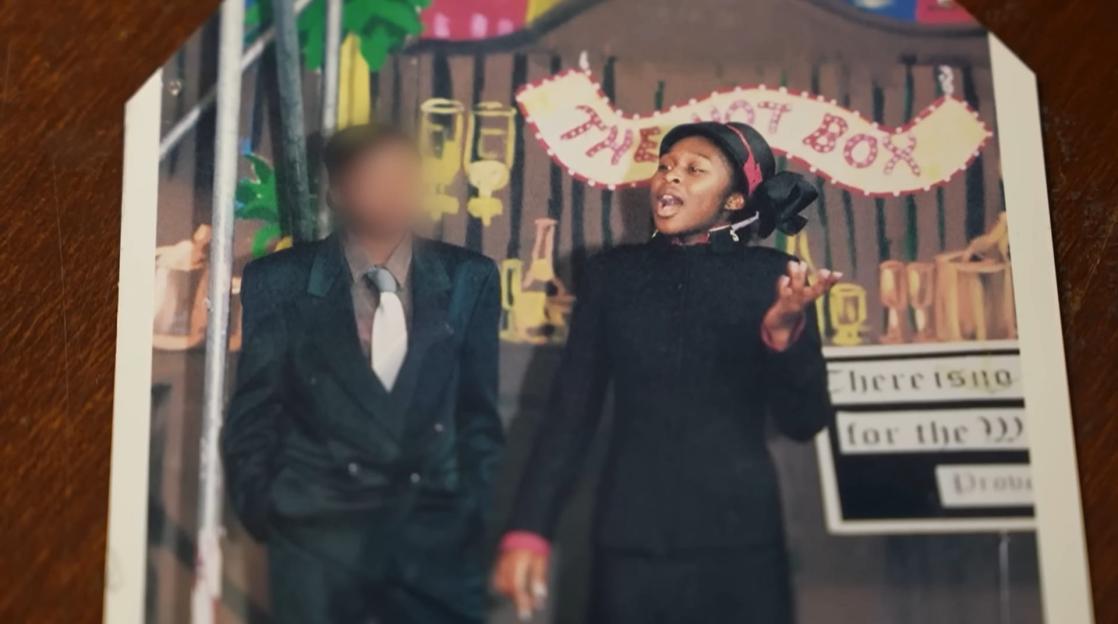 A young woman in a black suit and red headscarf sings on stage next to a blurred man in a suit, with a "The Hot Box" banner in the background.