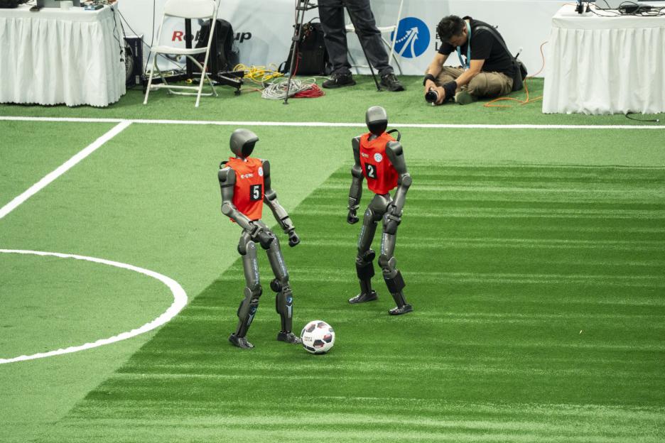 Two humanoid robots in red jerseys playing football on a green turf.