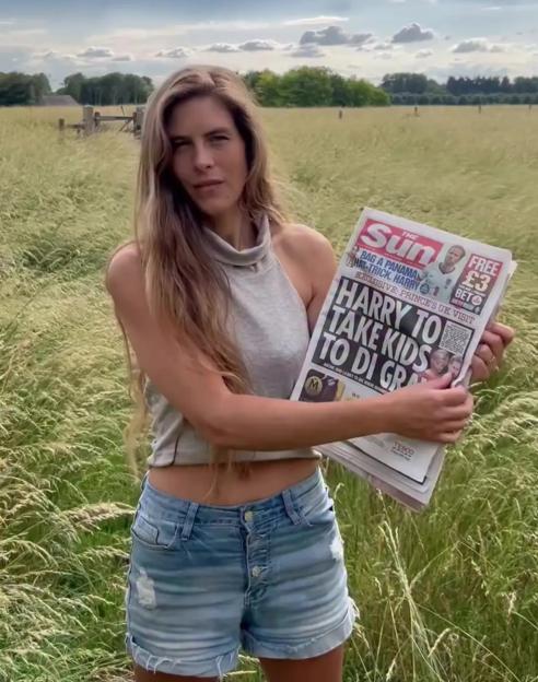 Hannah Bourne-Taylor holding up The Sun newspaper with the headline "HARRY TO TAKE KIDS TO DI GRA" while standing in a grassy field.