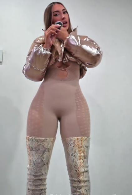 A woman in a form-fitting nude bodysuit with a cut-out on the chest, a cropped shiny gold puffer jacket, and snakeskin print thigh-high boots holds a small object to her mouth.