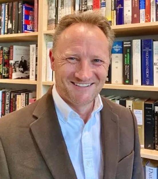 A man in a brown jacket and white collared shirt smiles in front of a bookshelf.
