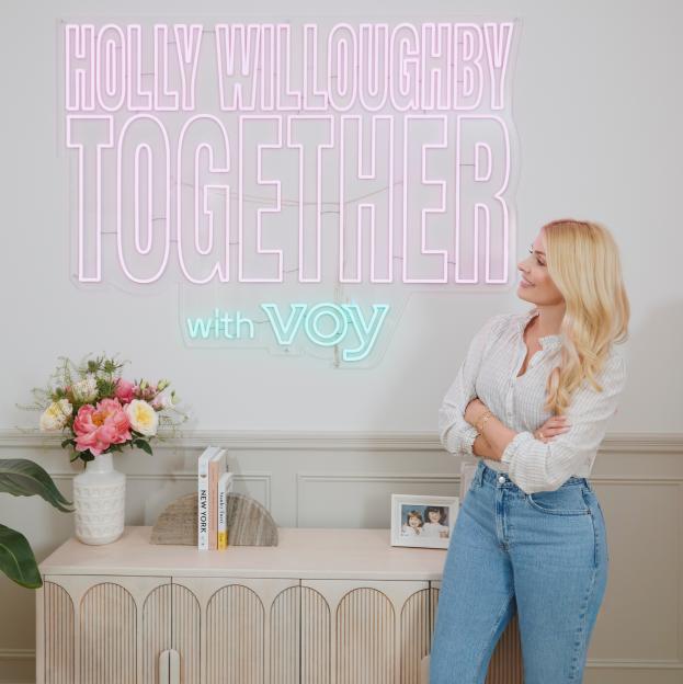 Holly Willoughby stands next to a pink and teal neon sign that reads "HOLLY WILLOUGHBY TOGETHER with VOY".