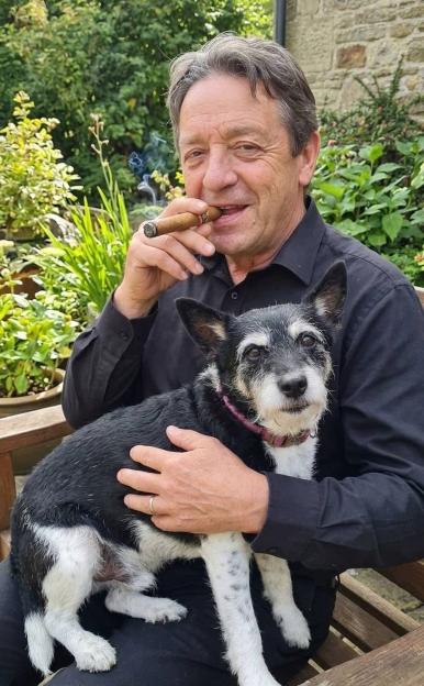 Francis Owens sitting on a wooden bench, holding a black and white terrier, with a cigar in his mouth, smiling.