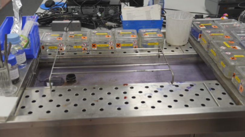 Lab equipment on a steel bench, including clear containers labeled with hazard symbols and a trough, suggesting chemical work.