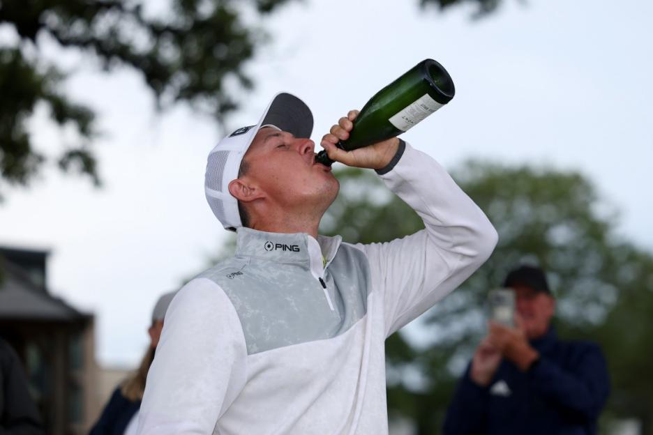 A golfer in a white and grey shirt and cap drinks from a green bottle.