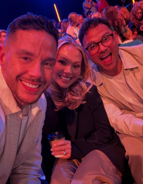 A man and a woman in the foreground smiling for a photo, with another man in glasses smiling with his mouth open to their right, all three in a crowd.