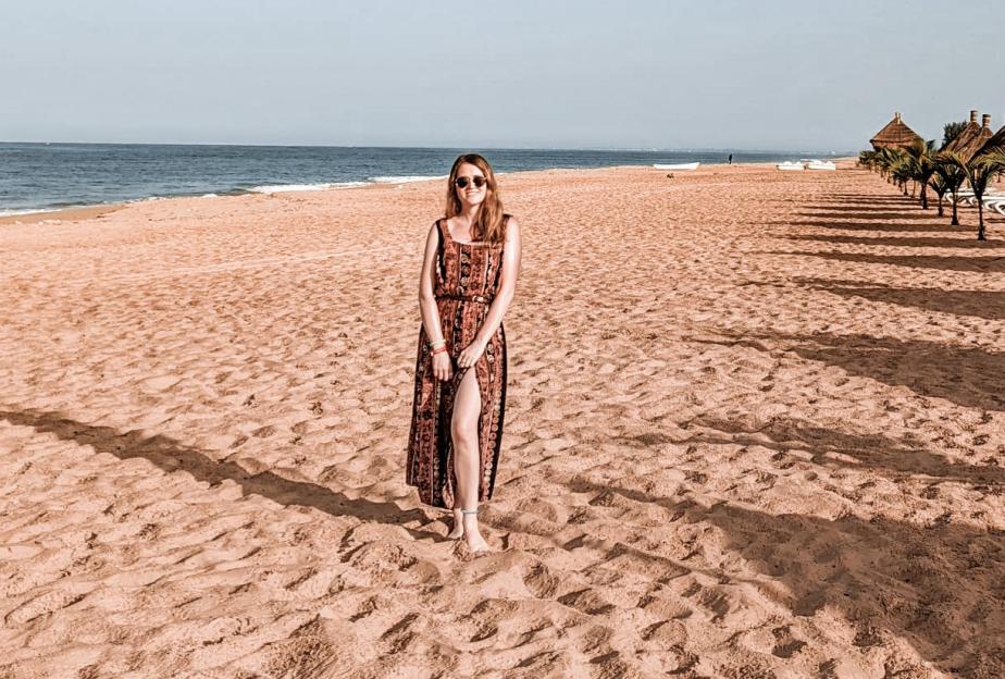 Woman in a dress and sunglasses standing barefoot on a sandy beach.