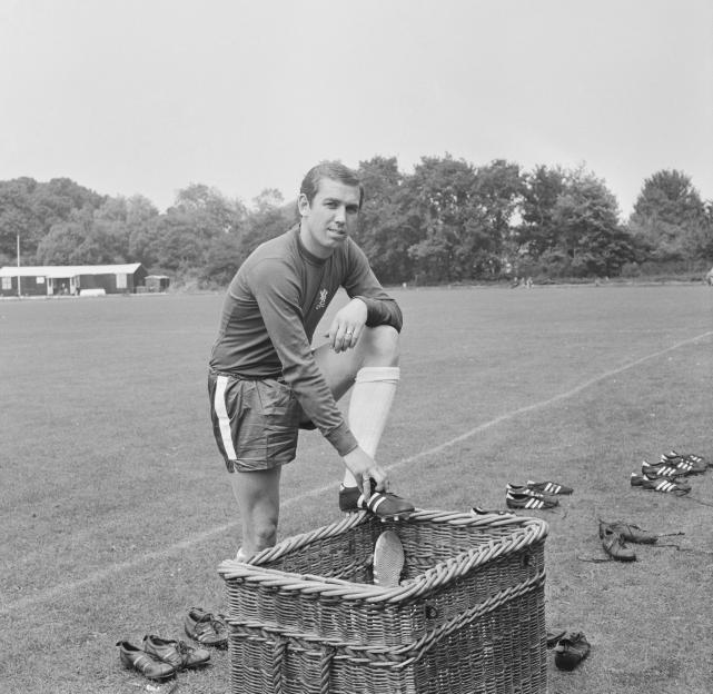 Marvin Hinton, former Chelsea defender, laces his boots with his foot on a wicker basket on a football pitch.
