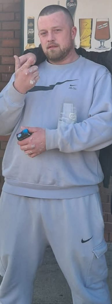 Astron Inman, a man with a beard and buzz cut, stands wearing a light grey Nike sweatshirt and sweatpants, his hands covered in white residue.
