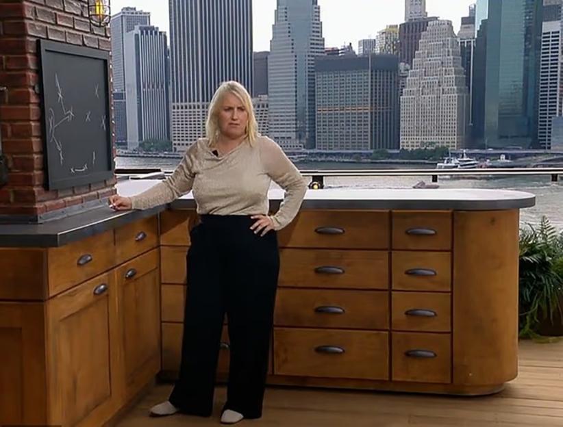 Woman in a shimmery top and black pants standing by a counter with a city skyline and water in the background.
