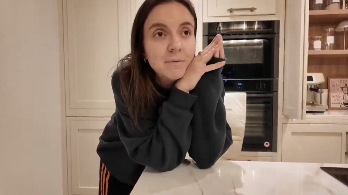 A woman with brown hair leans on a kitchen counter, looking away thoughtfully.