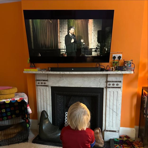 A child in a red shirt watching Alfie Brown perform stand-up comedy on TV.