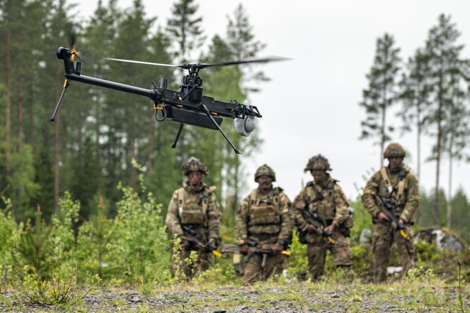 A Ghost drone with a rotating propeller and camera in the foreground, with four British Army soldiers in camouflage and gear standing in the background.