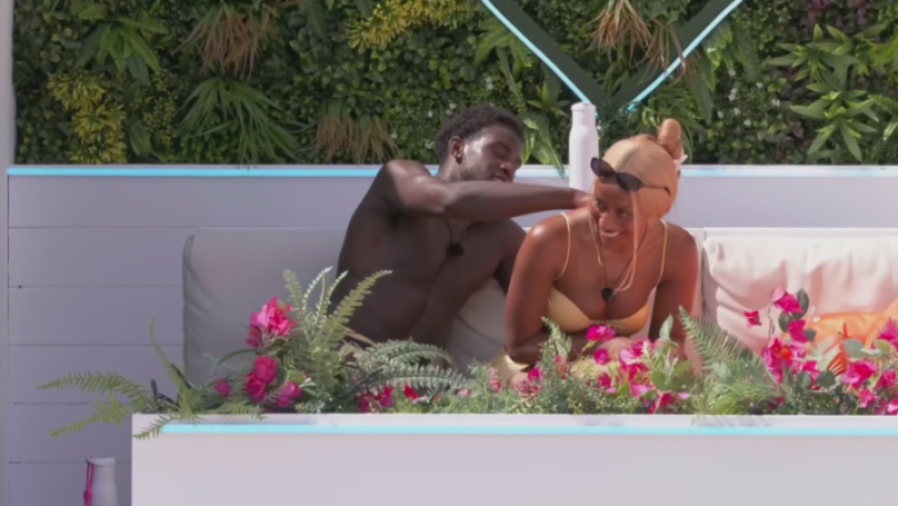 A man applies sunscreen to a woman's back while they sit on a white sofa.