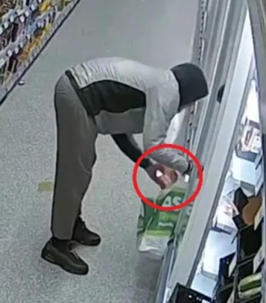 A person with a hoodie and gray clothing is bent over in a grocery store aisle, holding a bottle of wine and needles, indicated by a red circle.