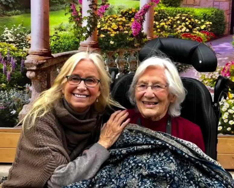 Two women, one younger and one older, sit together smiling in front of a floral backdrop.