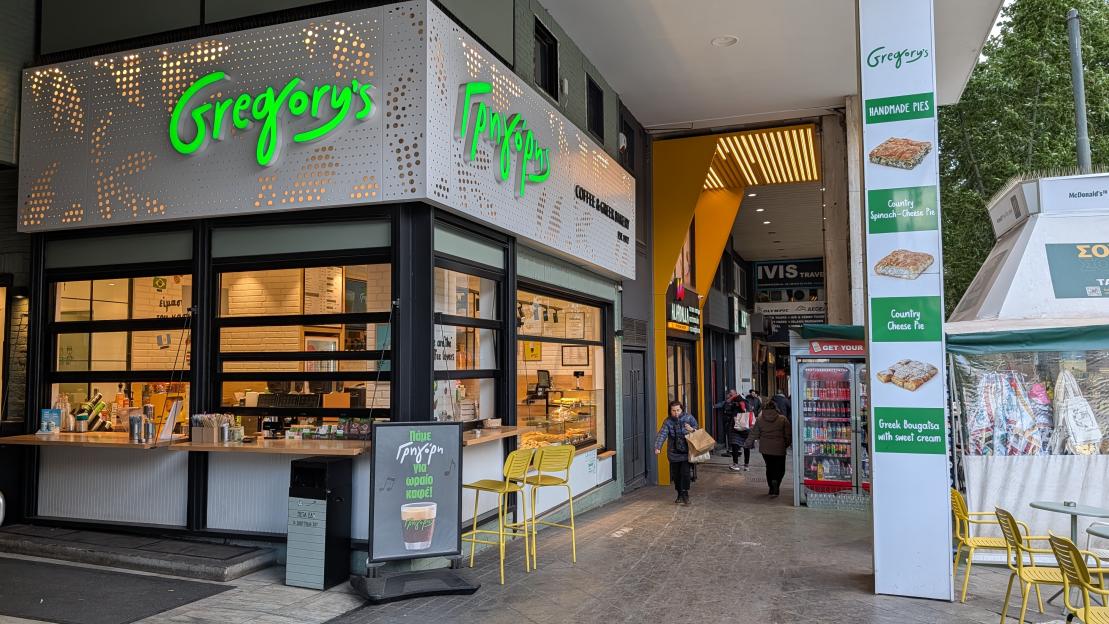 Exterior view of a Gregory's bakery and coffee shop, featuring its vibrant green sign, an open counter, and an advertisement for Greek pastries, with people walking by.