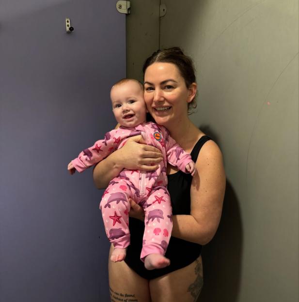 A woman in a black swimsuit holding a baby in a pink, dolphin-patterned swimsuit.