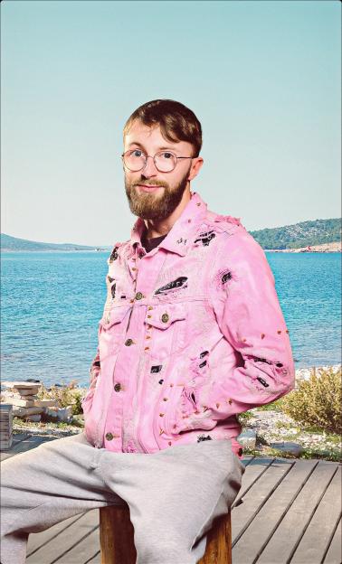 Man with a beard and glasses wearing a ripped pink denim jacket and gray sweatpants, sitting on a wooden bench with a blue ocean and island in the background.