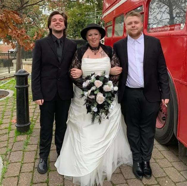 Samantha Fairbairn in a white wedding gown, black lace jacket and hat, holding a bouquet, stands between her two sons, both dressed in black suits, in front of a red double-decker bus.