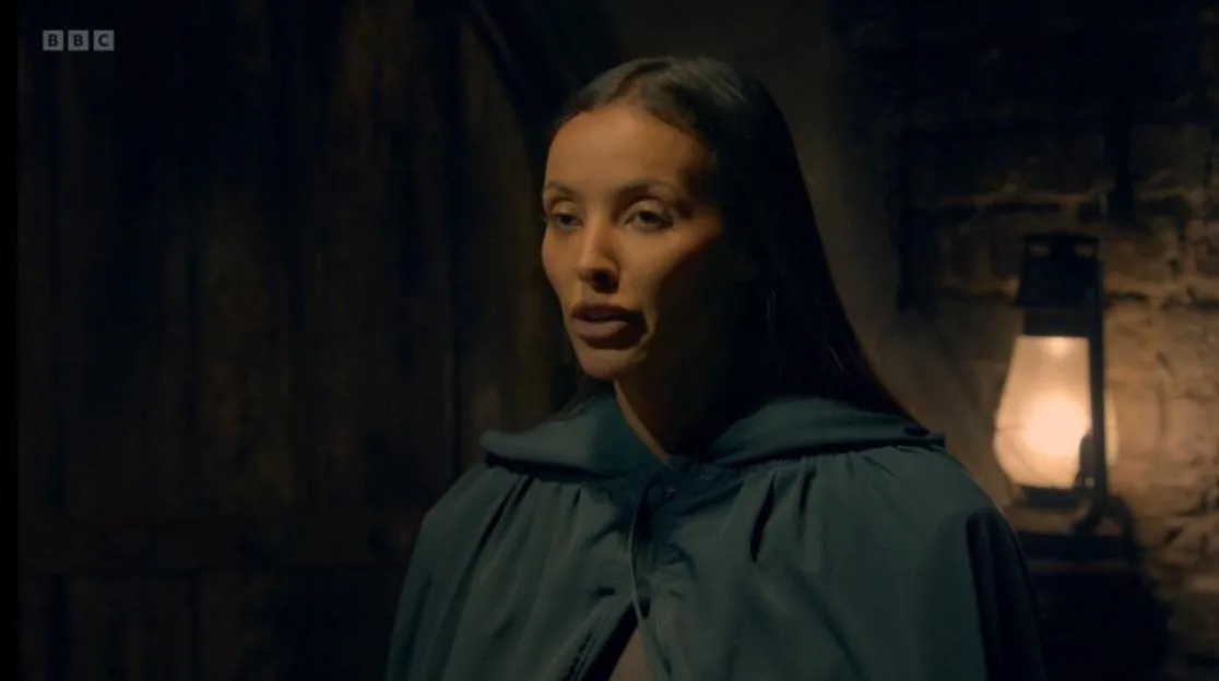 A woman with long dark hair wearing a dark cloak in a dimly lit room.