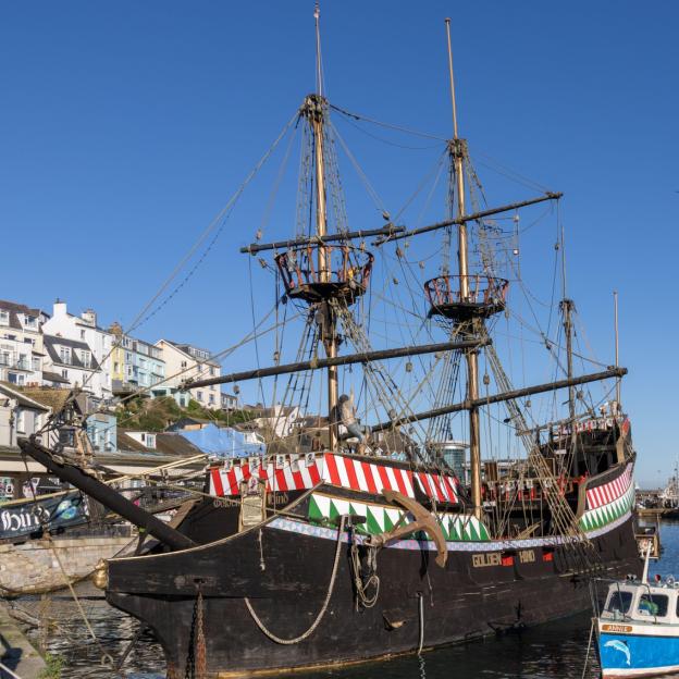 A replica of the Golden Hind docked in a harbor, with houses visible on the hillside in the background.
