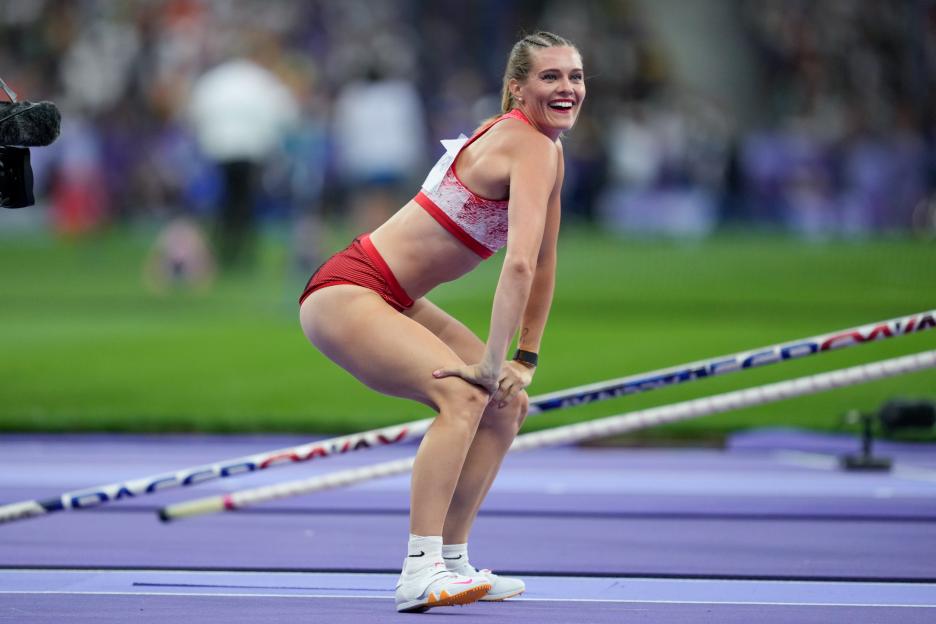 A female Olympic athlete in red and white sportswear smiles and laughs while bent over with her hands on her knees on a purple track.