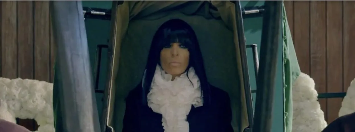 Claudia Winkleman stands inside an open coffin, wearing a dark coat and a large white ruffled collar.