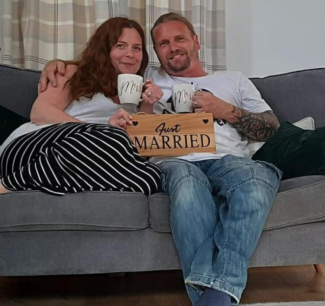 Married couple Chris and Kayleigh Coll sitting on a couch, holding "Mr" and "Mrs" mugs and a "Just Married" sign.