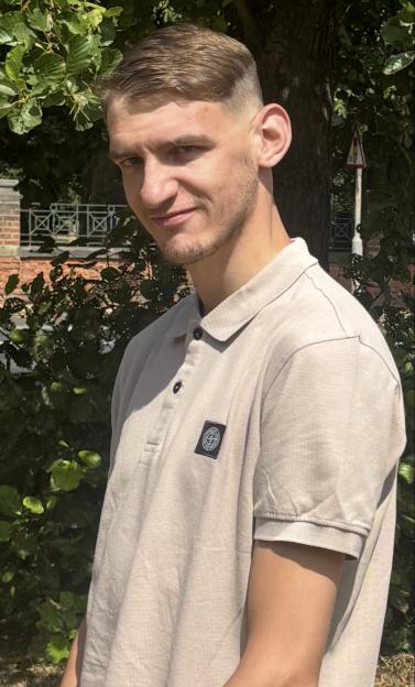 A young man with light brown hair and a short beard wears a light-colored polo shirt, looking at the camera with a subtle smirk.