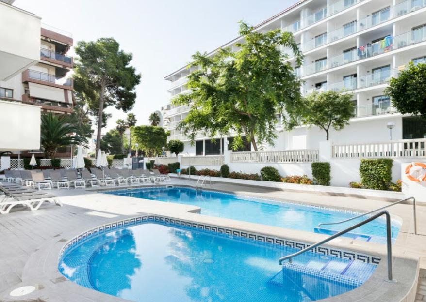 An outdoor swimming pool area with lounge chairs and a multi-story hotel building in the background.