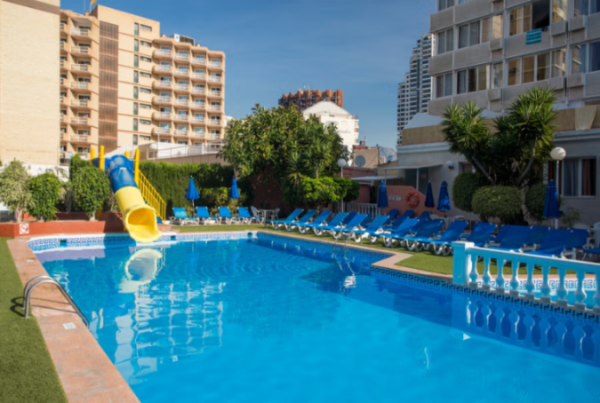 Swimming pool area with a water slide, blue lounge chairs, and hotel buildings.
